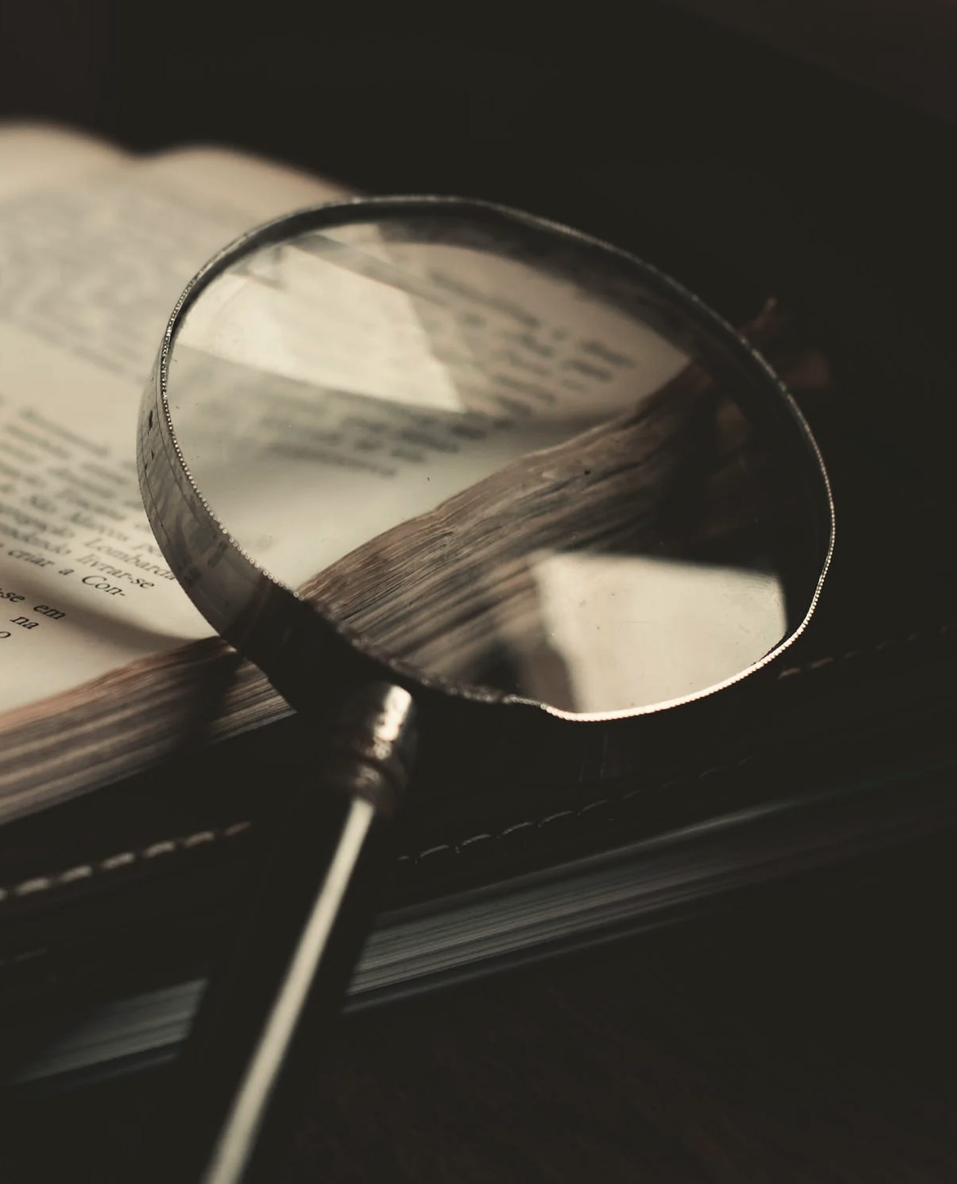 Magnifying glass on an open book with visible text, creating a vintage, investigative mood. Soft lighting highlights the page details.