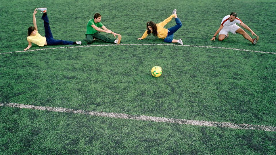 Four people doing leg and hamstring stretches on green artificial turf, with a yellow soccer ball in the middle of the field.
