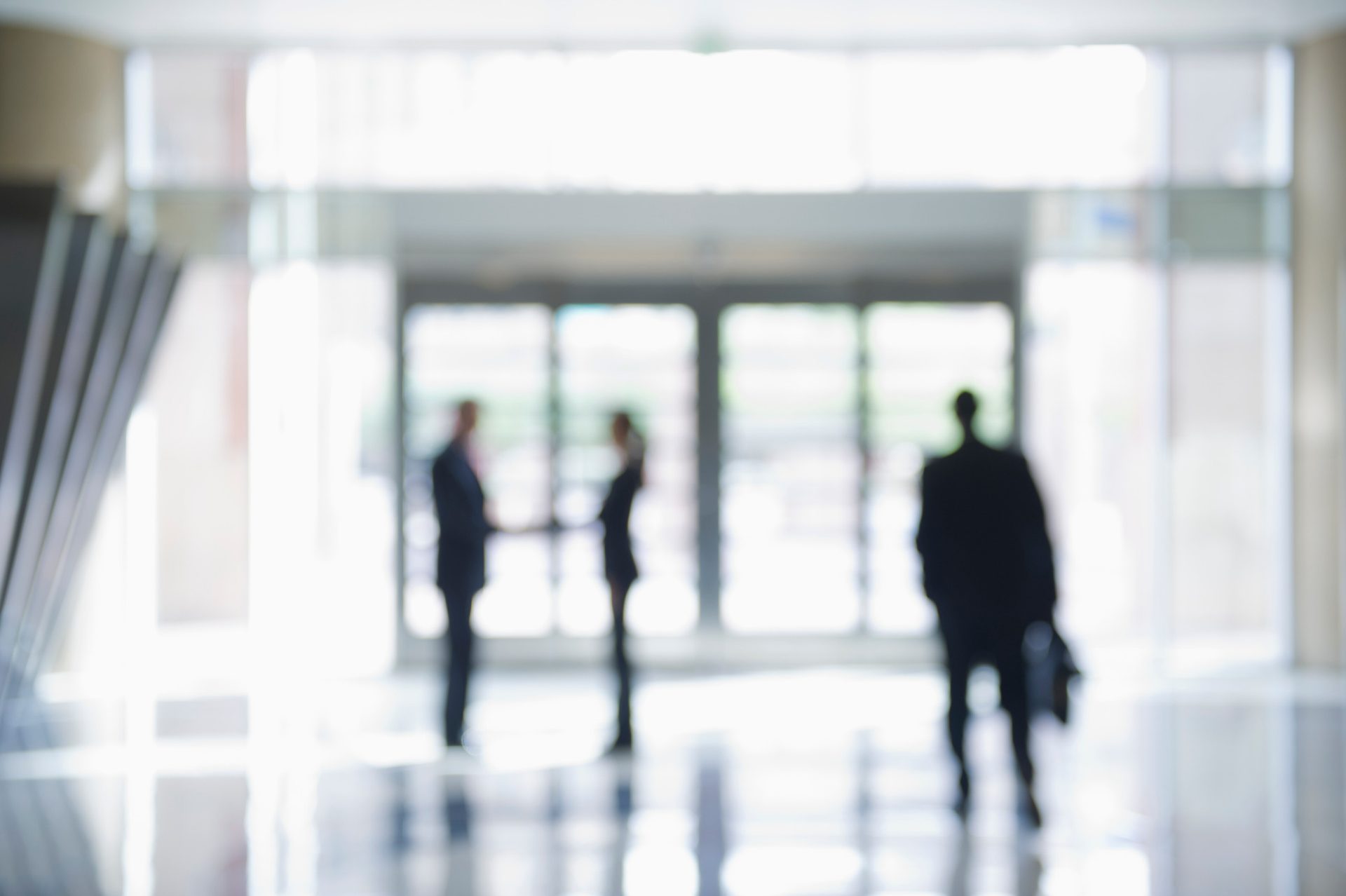 Blurred silhouettes of two people shaking hands and a third person walking past in a bright office lobby.