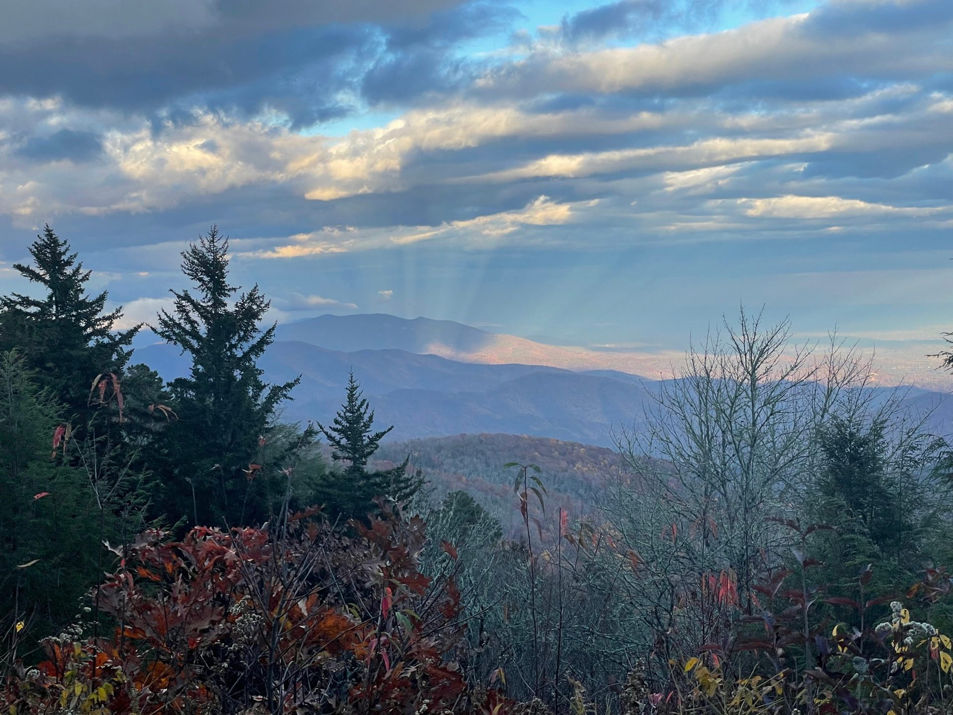 Rays of sunlight breaking through clouds above forested mountain ridges in autumn.