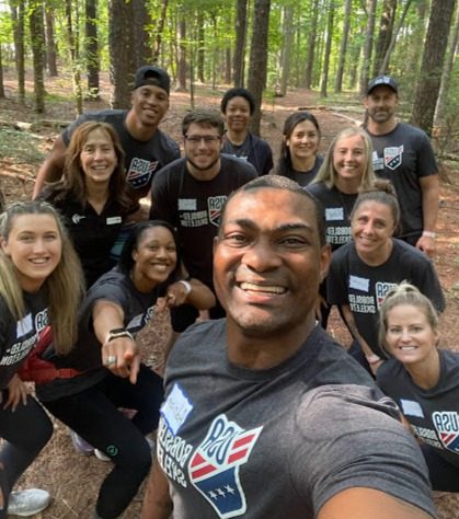 A group of smiling people in matching dark T-shirts posing close together for a selfie in a pine woodland.