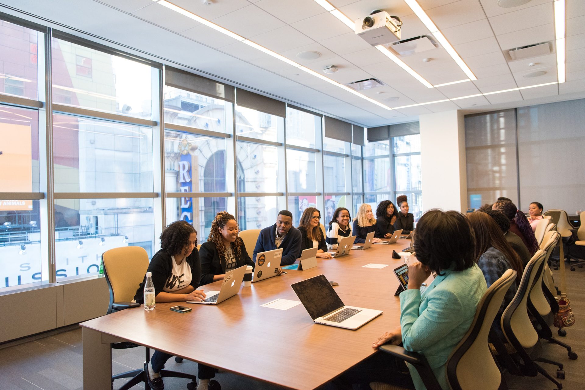 People seated around a long wooden table in a bright conference room, most of them working on laptops.